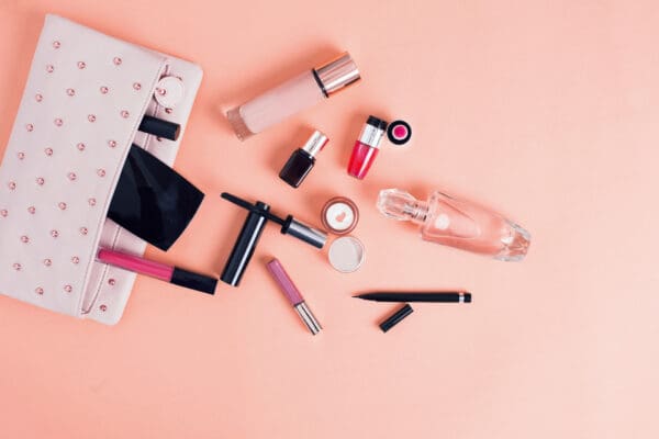 A Care Package Quarterly makeup bag filled with lipsticks, nail polish, mascara, a perfume bottle, and eyeliner pencils is artfully arranged against a peach background.