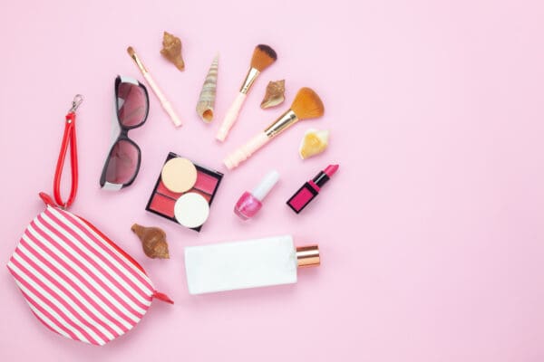 A pink background featuring scattered cosmetics, brushes, sunglasses, seashells, and a striped makeup bag from the Care Package Quarterly collection.