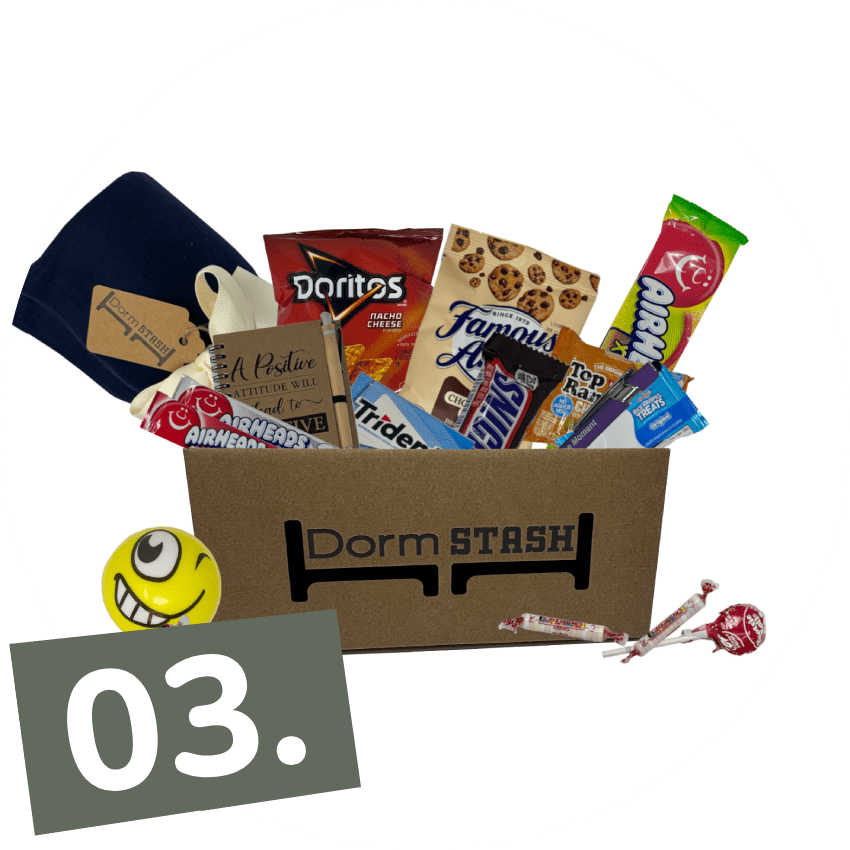 A Dorm Stash box filled with snacks like Doritos, Famous Amos cookies, Airheads, and other treats, along with a blanket and a smiley face stress ball. The box is labeled "03.