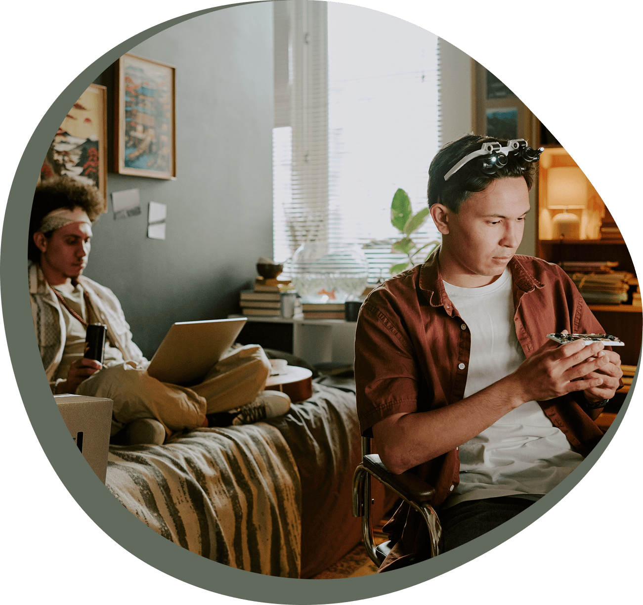 25 Two young adults in a cozy room; one sits on a bed with a laptop and drink, while the other sits at a desk, looking at something in their hands.